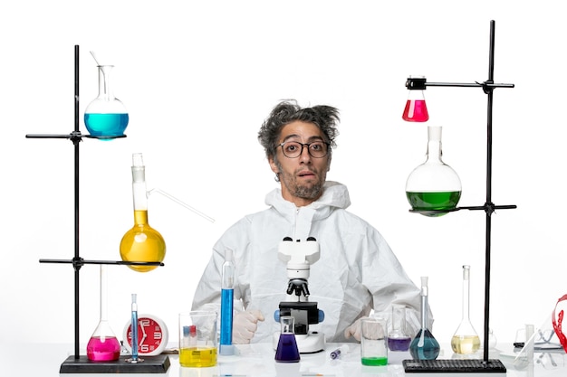 Front view crazy male scientist in special protective suit sitting around table with solutions on light white background lab disease covid- science virus