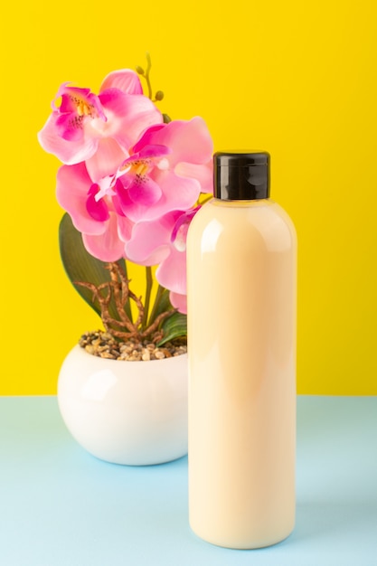 A front view cream colored bottle plastic shampoo can with black cap isolated along with flowers on the yellow-iced-blue background cosmetics beauty hair