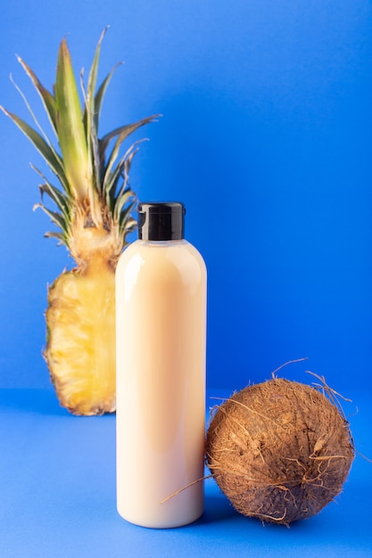 A front view cream colored bottle plastic shampoo can with black cap isolated along with sliced pineapple and coconut on the blue background cosmetics beauty hair