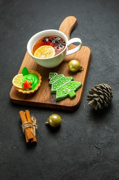 Front view of a cup of black tea xsmas accessories conifer cone and cinnamon limes on a wooden cutting board on black background