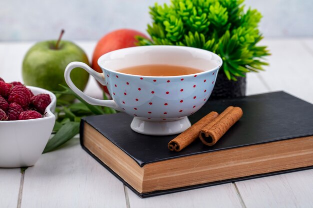 Front view of cup of tea on a book with cinnamon raspberries and apples on a white surface