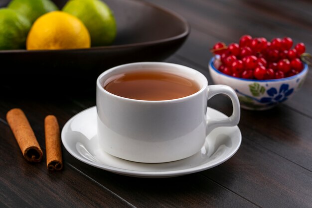 Front view of cup of tea with cinnamon and red currants in a bowl on a wooden surface