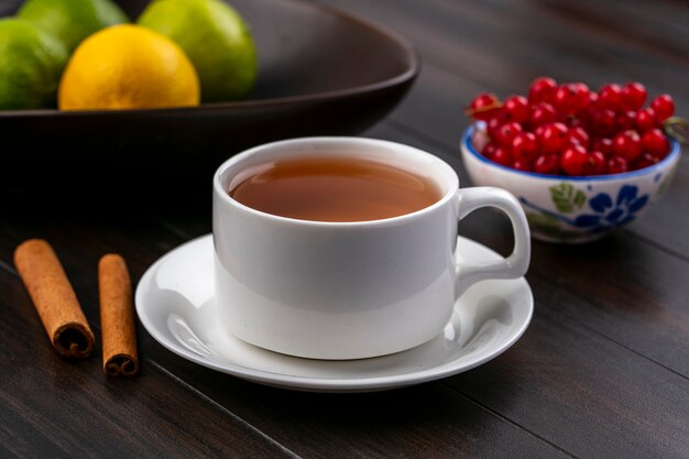 Вид спереди чашки чая с корицей и красной смородиной в миске на деревянной поверхности