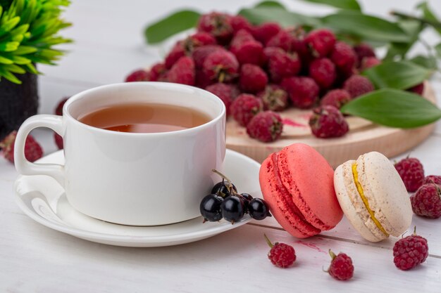 Front view of cup of tea with macaroon and raspberries on a white surface