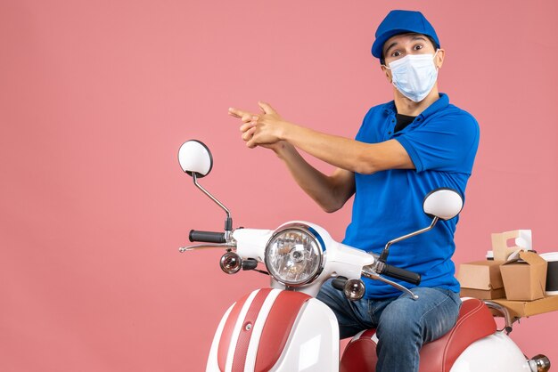 Front view of curious courier man in medical mask wearing hat sitting on scooter on pastel peach background