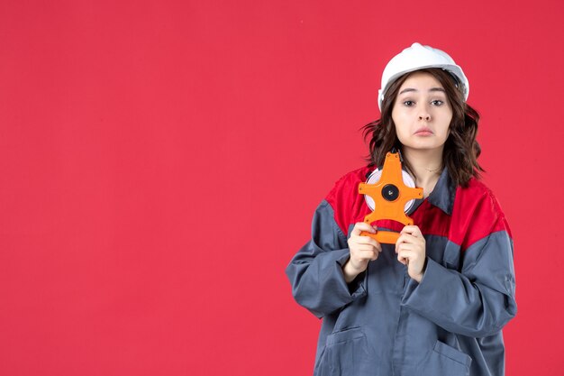 Front view of curious female architect in uniform with hard hat holding measuring tape on isolated red wall