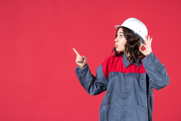 Front view of curious female builder in uniform with hard hat and making eyeglasses gesture pointing up on isolated red background