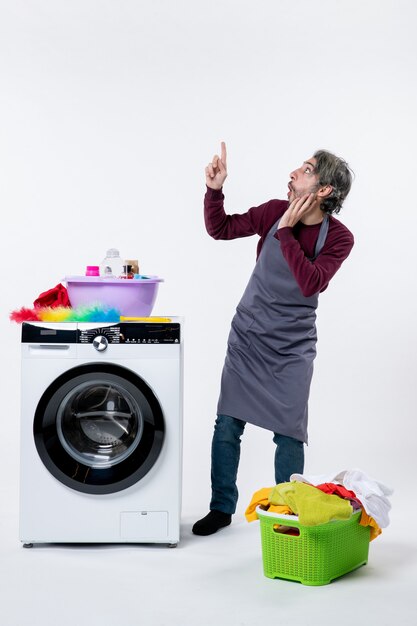 Front view curious housekeeper man standing near washer laundry basket on white background