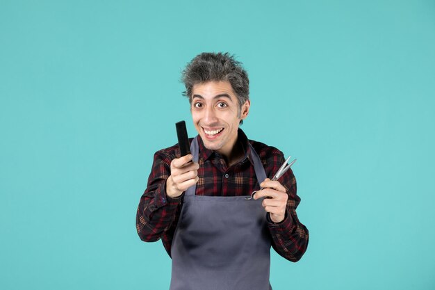 Front view of curious male hairdesser wearing gray apron and holding scissor comb on blue soft color background