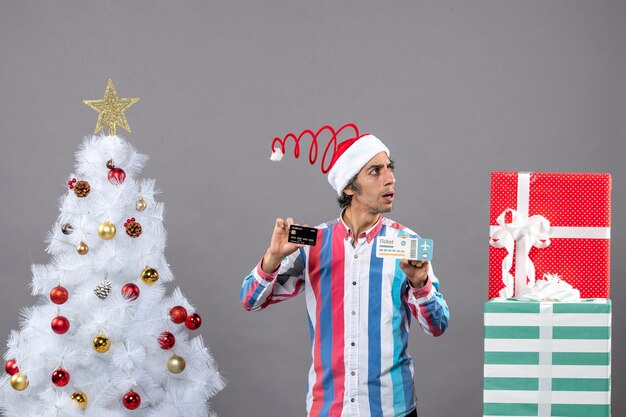 Front view curious man looking at right holding card and travel ticket around xmas tree and presents