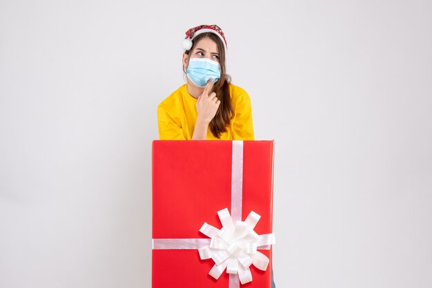 Front view curious xmas girl with santa hat standing behind big xmas gift