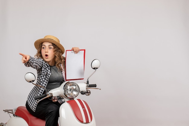 Free photo front view of curious young girl on moped holding up clipboard on grey wall
