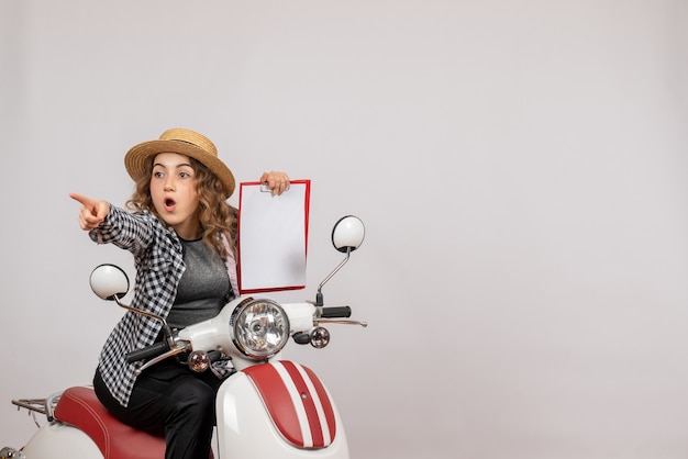 Front view of curious young girl on moped holding up clipboard on grey wall