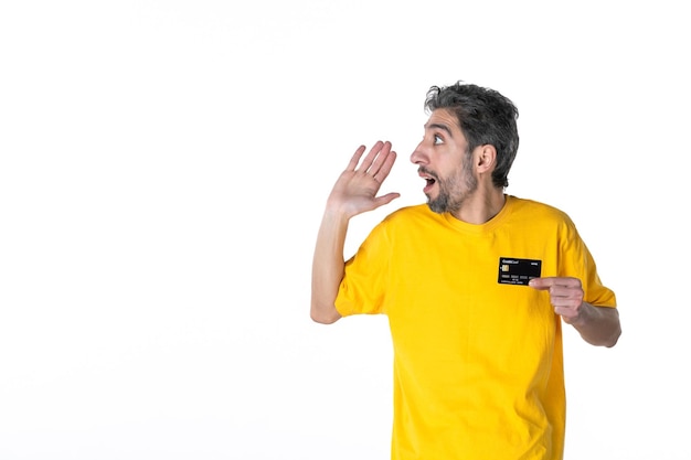 Front view of curious young male in yellow shirt and holding bank card and looking at something on the right side on white background