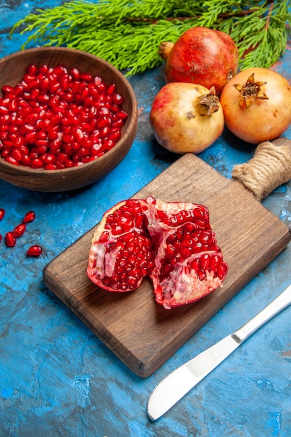 Front view a cut pomegranate and dinner knife on chopping board pomegranate seeds in bowl and pomegranates on blue background