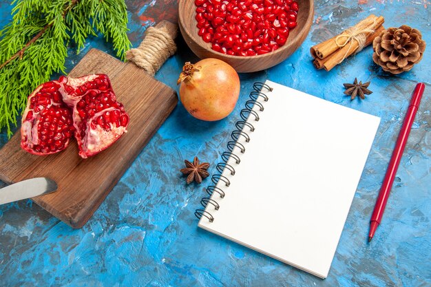 Front view a cut pomegranate and dinner knife on chopping board pomegranate seeds in bowl and pomegranates cinnamon anise seeds a notebook red pen on blue background