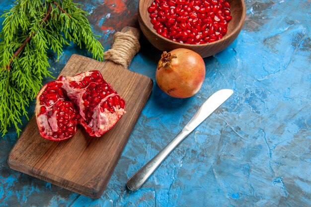 Front view a cut pomegranate and dinner knife on cutting board pomegranate seeds in bowl and pomegranate on blue background free space