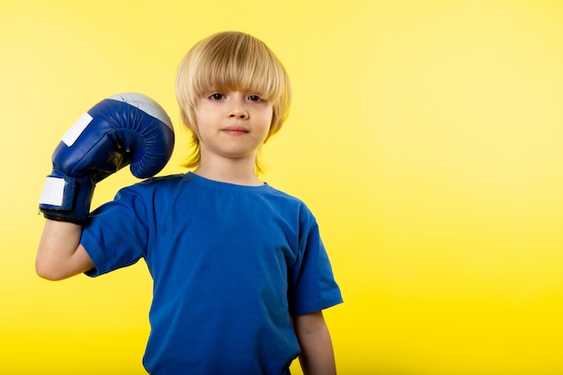 Free photo a front view cute blonde boy posing in blue t-shirt and blue glove on the yellow wall