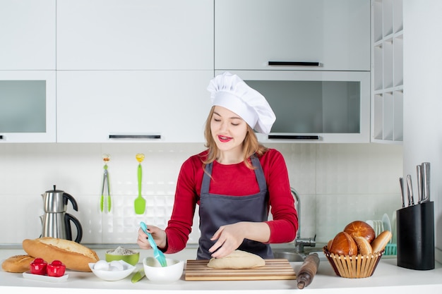 Front view cute blonde woman in cook hat and apron buttering bread in the kitchen