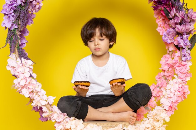 A front view cute boy in white t-shirt eating choco donuts on the yellow desk