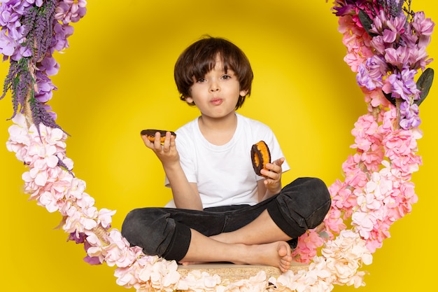 A front view cute boy in white t-shirt eating donuts on the flower made stand on the yellow space