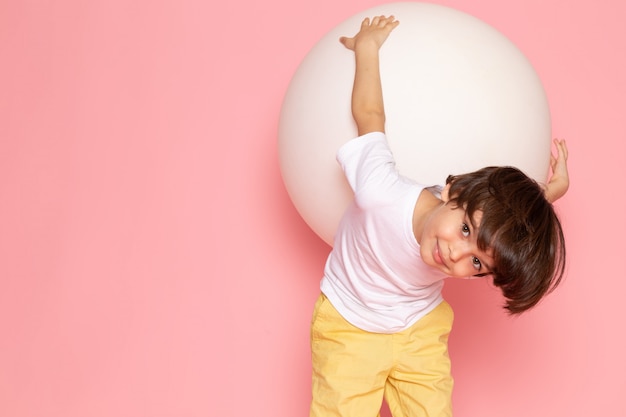 A front view cute boy in white t-shirt playing with white ball on the pink floor