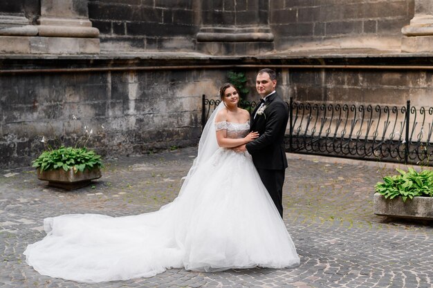 Front view of cute bride in wedding gown with long train and veil standing near groom holding his hand looking and posing at camera during wedding walk in ancient city