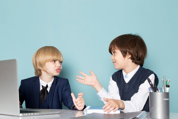 A front view cute business boy in blue classic suit posing in front of silver laptop along with other boy discussing working business work fashion