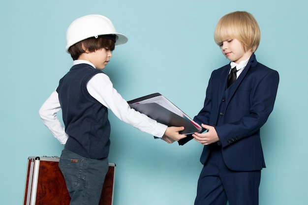 フォルダービジネス仕事ファッションを与える他の少年と一緒に茶色銀のスーツケースを持ってポーズをとって青いクラシックスーツの正面かわいいビジネスボーイ