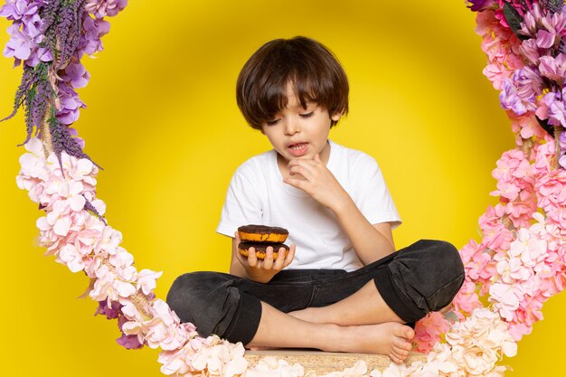 A front view cute child eating choco donuts in white t-shirt on the yellow floor