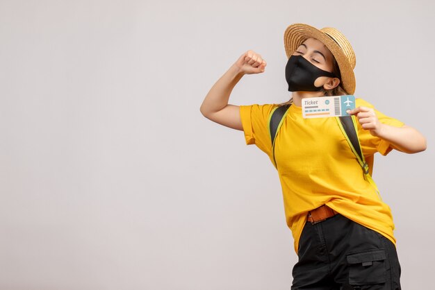 Front view of cute female traveller with backpack wearing black mask holding travel ticket on white wall