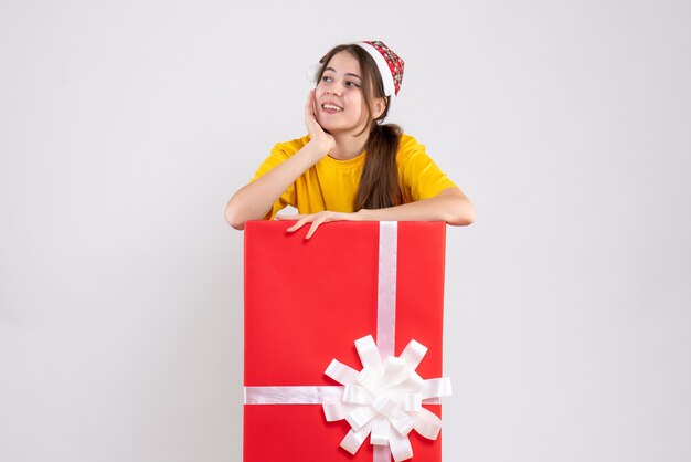 Вид спереди милая девушка в шляпе санта-клауса смотрит на что-то, стоящее за большим рождественским подарком