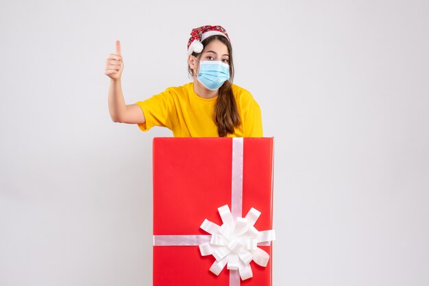 Front view cute girl with santa hat making thumb up sign standing behind big xmas gift