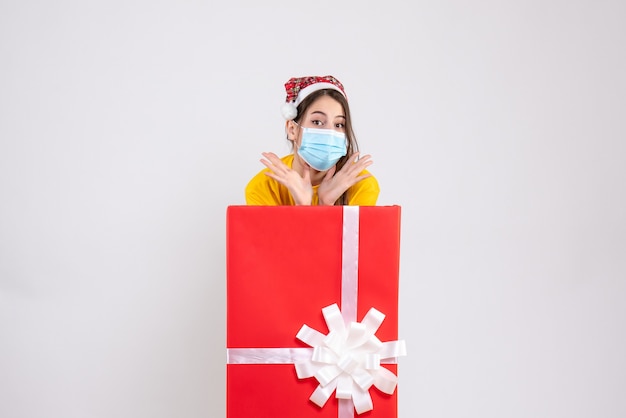 Front view cute girl with santa hat and mask standing behind big xmas gift