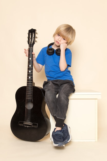 Вид спереди милый маленький мальчик в синей футболке с черными наушниками держит черную гитару