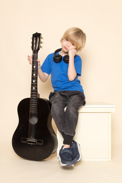 A front view cute little boy in blue t-shirt with black headphones holding black guitar