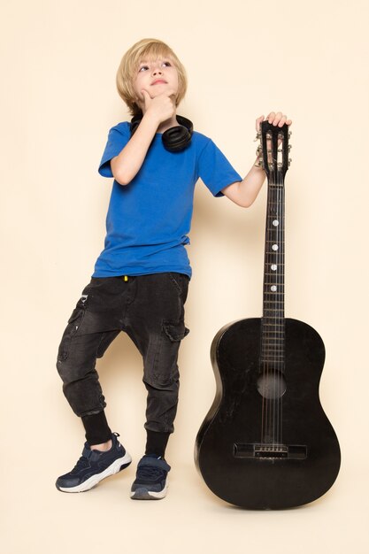 A front view cute little boy in blue t-shirt with black headphones holding black guitar