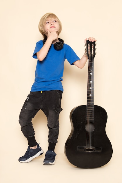 Вид спереди милый маленький мальчик в синей футболке с черными наушниками держит черную гитару