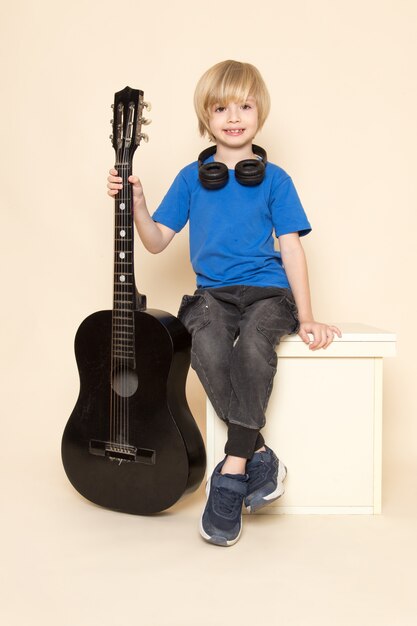 A front view cute little boy smiling in blue t-shirt with black headphones holding black guitar