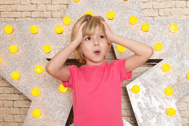 A front view cute little kid in pink t-shirt grey jeans on the star designed yellow stand and light background