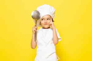 Free photo a front view cute little kid in white cook suit and white cook cap holding big spoon on the yellow wall child cook kitchen food