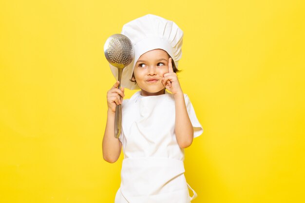 A front view cute little kid in white cook suit and white cook cap holding big spoon on the yellow wall child cook kitchen food