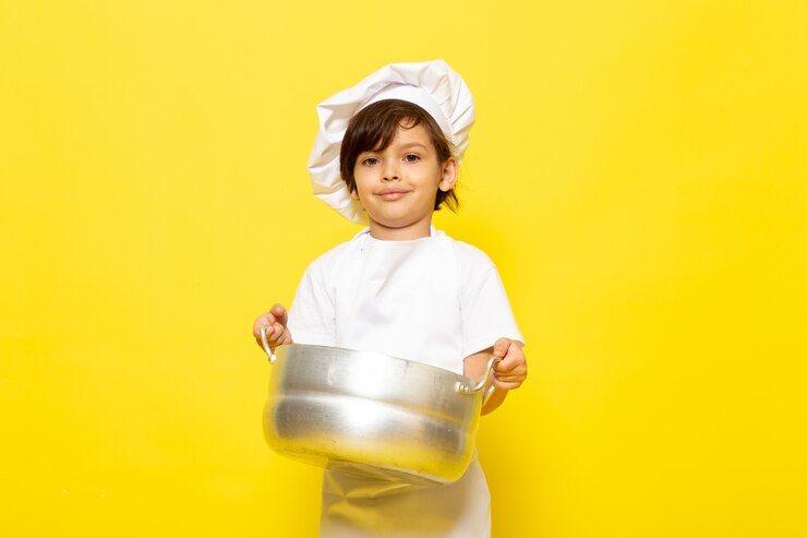 Free Photo a front view cute little kid in white cook suit and white cook cap holding silver pan smiling on the yellow wall child cook kitchen food