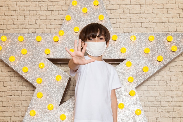 A front view cute little kid in white t-shirt dark jeans white sterile mask on the star designed yellow stand and light background