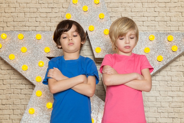 A front view cute little kids in blue and pink t-shirts dark and grey jeans on the star designed yellow stand and light background