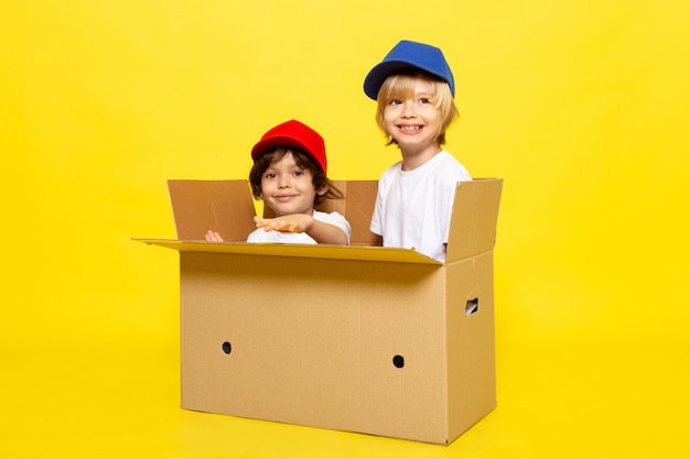 Вид спереди милые маленькие дети в белых футболках красные и синие шапки, улыбаясь внутри коричневой коробке на желтой стене