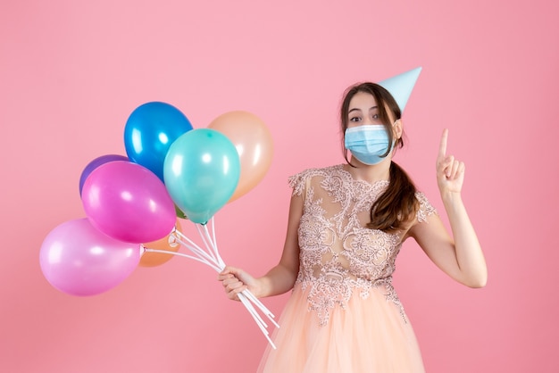 Front view cute party girl with party cap holding colorful balloons pointing with finger up