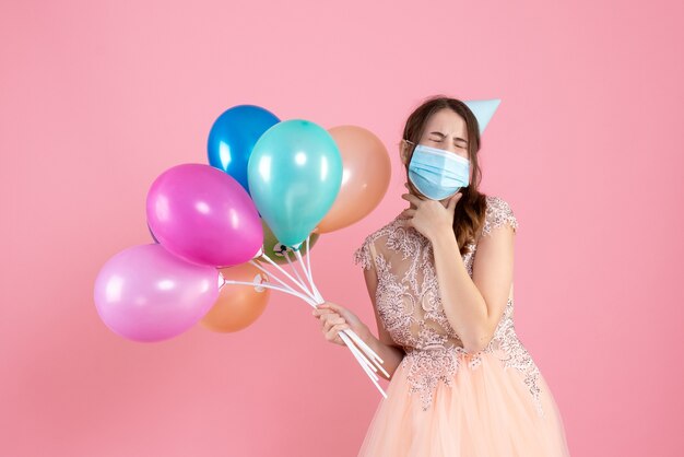Front view cute party girl with party cap and medical mask closing her eyes holding colorful balloons