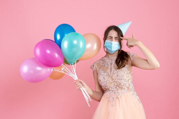 Front view cute party girl with party cap and medical mask holding colorful balloons putting finger gun to her temple