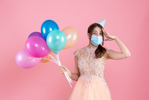Front view cute party girl with party cap and medical mask holding colorful balloons putting hand to her forehead